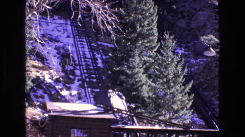 1973 Colorado home-movie frame looking across a rustic lodge roof toward evergreen trees and a snow-dusted inclined railway on the mountain slope.