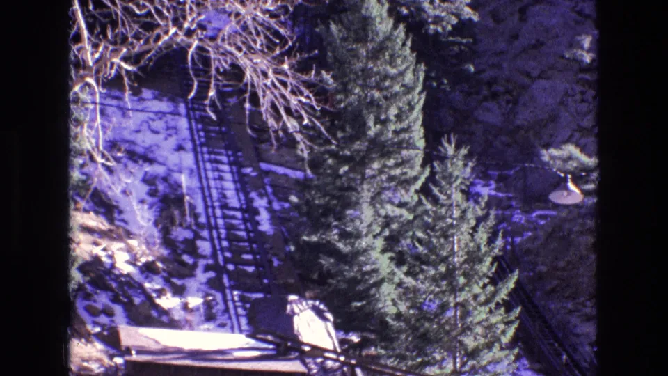 1973 Colorado home-movie frame with a steep rail line, winter snow, dense evergreen trees, bare branches, and a small object or visitor area near the lower right.