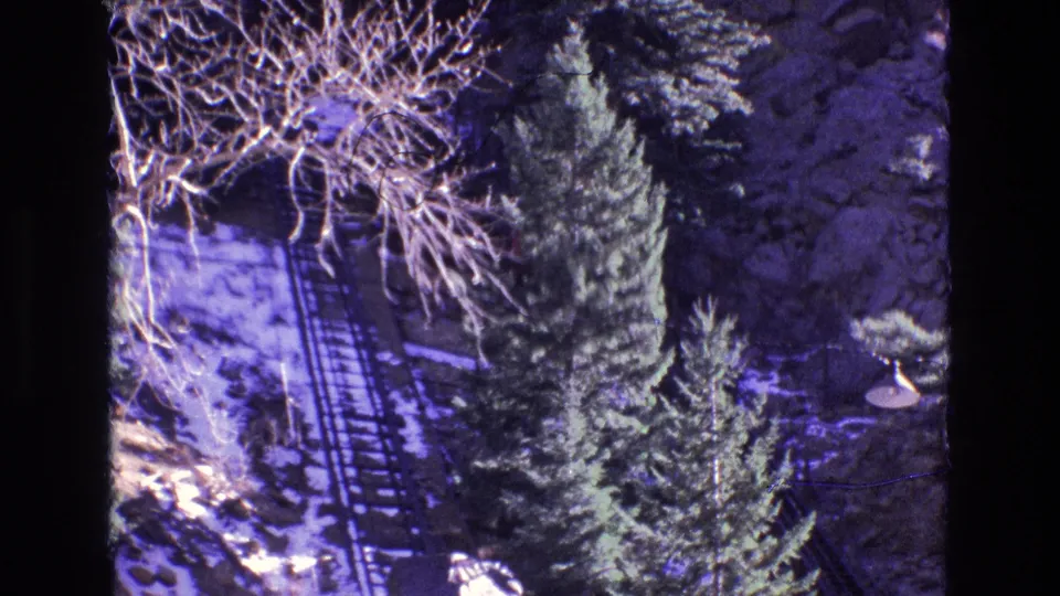 Colorado 8mm frame from 1973 looking down over an incline railway, with snow, pines, bare limbs, and a lower structure or platform near the track base.