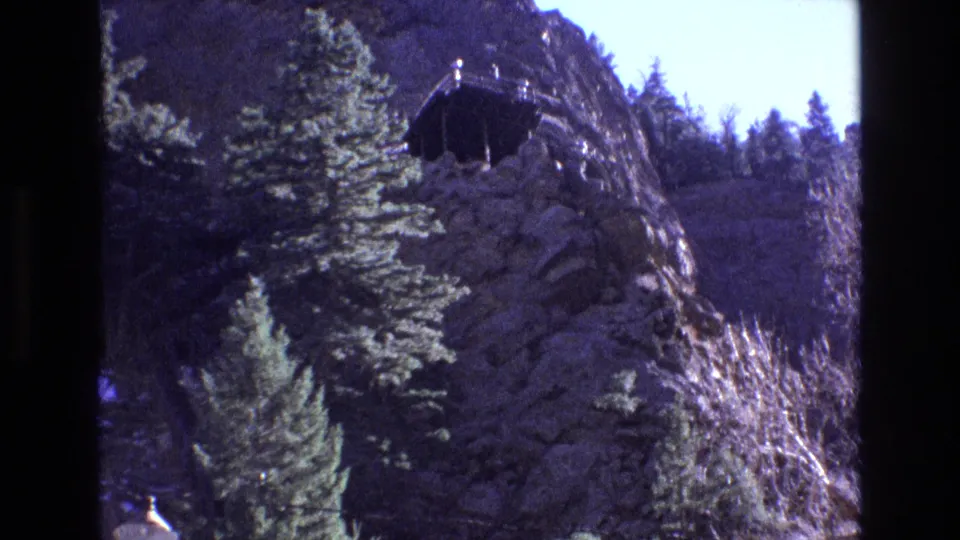 1973 Colorado home-movie frame showing a steep rocky hillside with evergreen trees and a small dark structure or cable-car element near the upper slope.