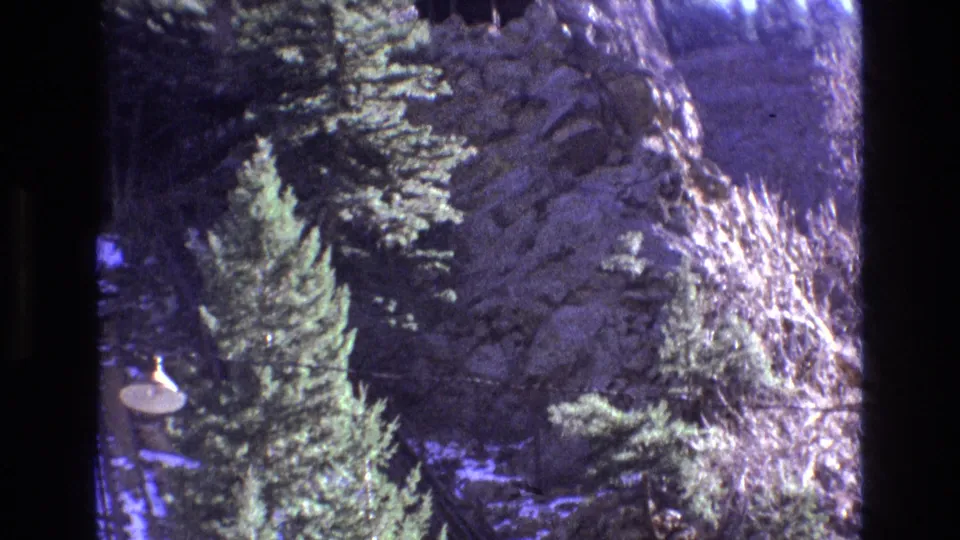 1973 Colorado 8mm frame with evergreen trees, an outdoor lamp, rocky terrain, and diagonal rail or cable-like lines running along the steep slope.