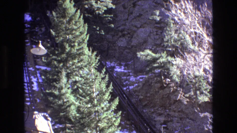 1973 Colorado home-movie frame showing evergreen trees, rocky hillside, outdoor lamps, diagonal transport lines, and a small person-like figure near the lower edge.