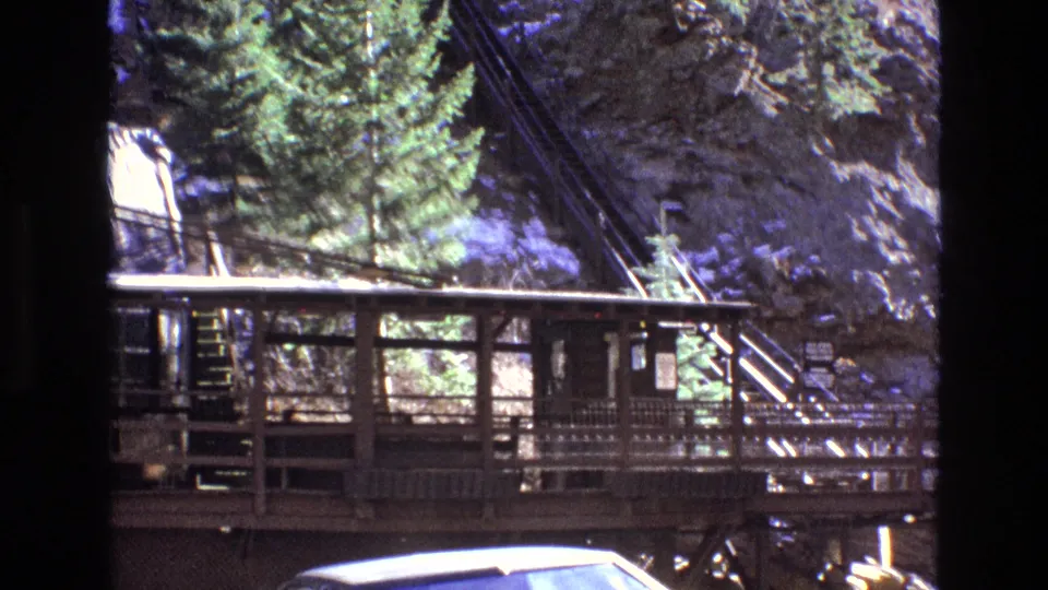 1973 Colorado 8mm frame showing a mountain railway platform above a yellow vintage American car, with evergreen trees, stairs, and rock formations in the background.