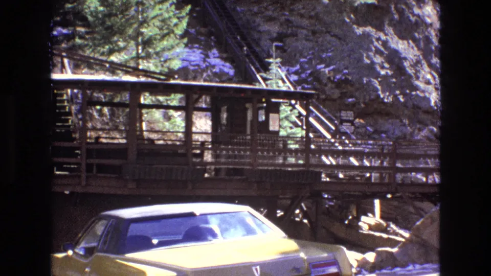 1973 Colorado home-movie frame of a yellow vintage car parked beneath a wooden mountain railway station platform, with stairs, railings, trees, and rocky terrain visible.