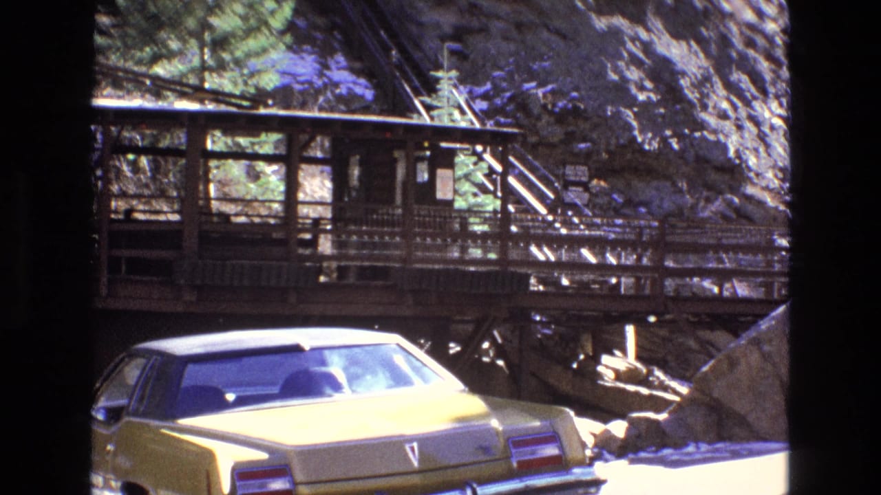 1973 Colorado home-movie frame showing a yellow sedan near a rustic wooden walkway built against a rocky, snow-dusted mountainside.