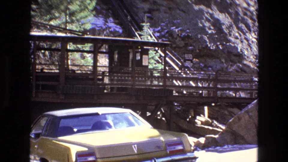 1973 Colorado home-movie frame showing a yellow sedan near a rustic wooden walkway built against a rocky, snow-dusted mountainside.