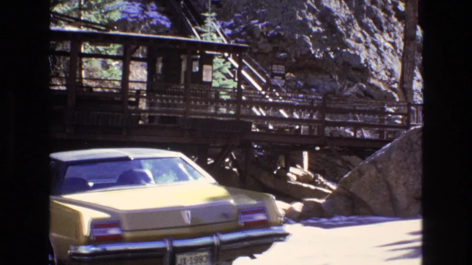 Yellow 1970s sedan seen from the rear beneath a raised wooden walkway and railings in a Colorado mountain park setting.