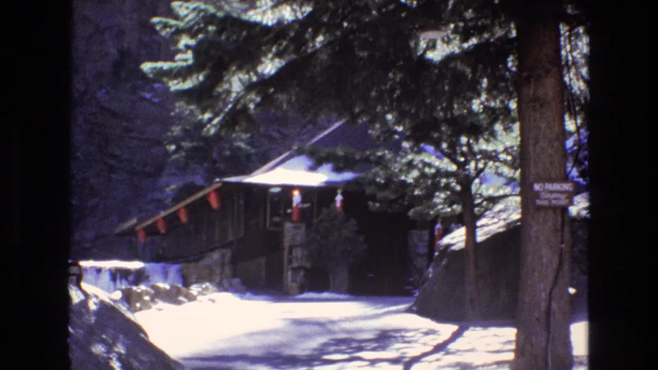 1973 Colorado 8mm frame of a snow-lined path, small rustic building, evergreen trees, hanging red markers, and a no-parking sign attached to a tree.