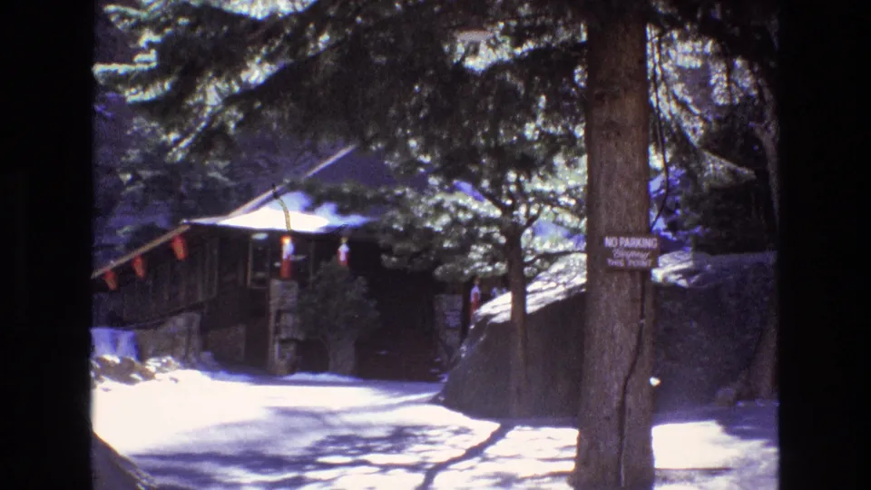 Colorado home-movie frame showing a rustic mountain park entrance area with snow, trees, a small building, and a no-parking sign visible on a trunk.