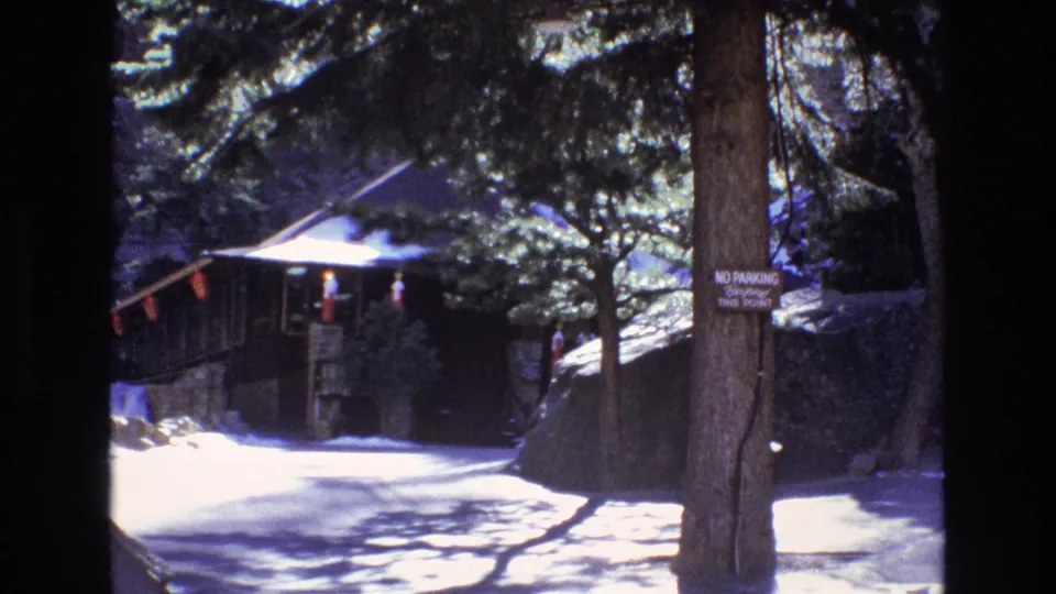 1973 Colorado travel footage frame with a tree-lined path, rustic building, bright sun patches, and a no-parking notice posted on a trunk.