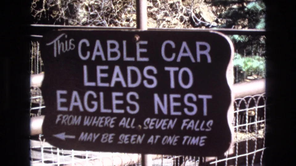 1973 Colorado home-movie frame showing a dark directional sign with white lettering for a cable car to Eagles Nest, with wire fencing and sunlit outdoor background visible.