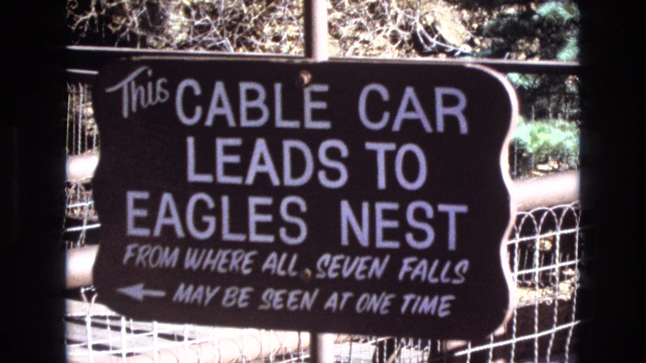 Dark cable car sign with white lettering behind a wire fence.