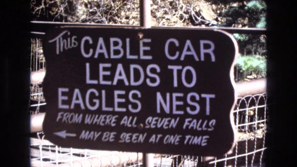 1973 Colorado frame of the Eagles Nest cable car sign, with the message about seeing all seven falls and a left-pointing arrow visible behind wire fencing.