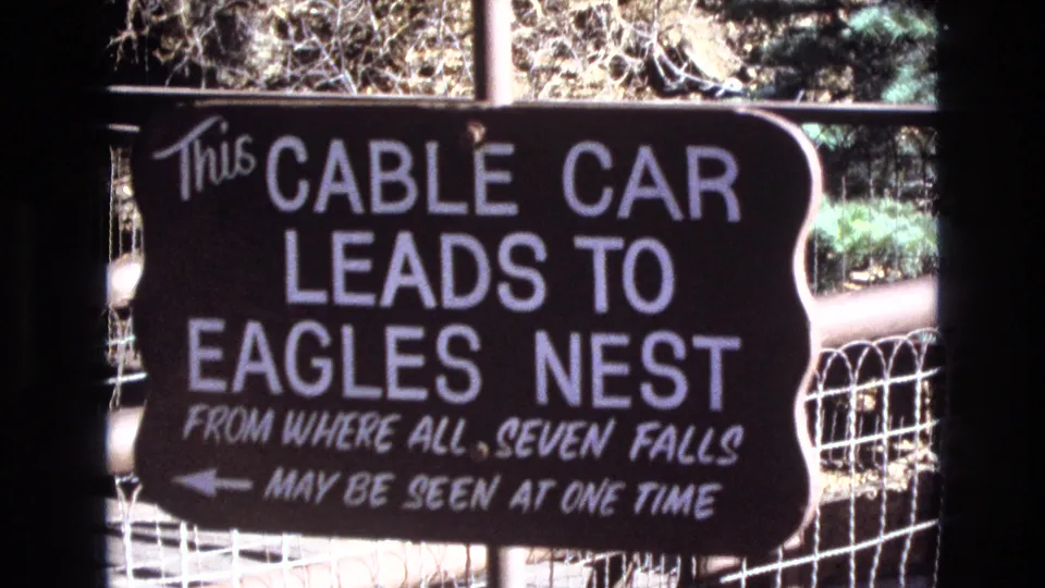 1973 Colorado 8mm frame showing a close visitor-shot view of the cable car sign, with the large Eagles Nest text, smaller Seven Falls wording, and fence lines behind it.