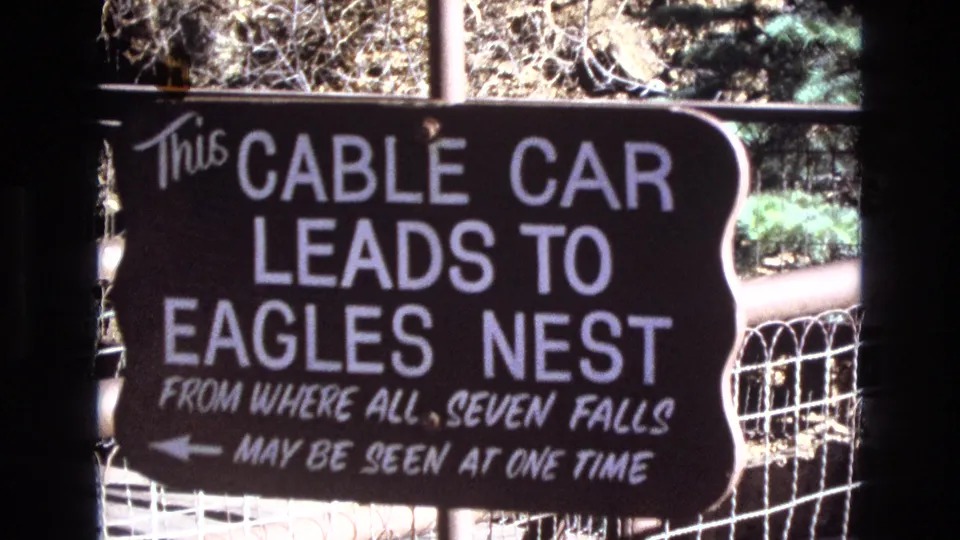 Final 1973 Colorado frame holding on the dark cable car sign for Eagles Nest and Seven Falls, with white painted text, a left arrow, wire fence, and sunlit vegetation in the background.