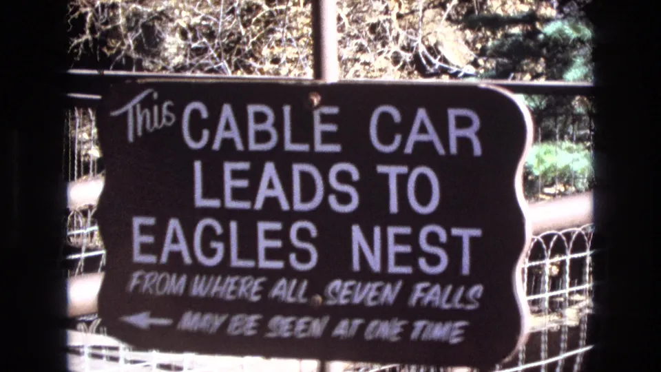 1973 Colorado 8mm frame showing a hand-painted sign that reads that a cable car leads to Eagles Nest, with wording about seven falls and trees behind it.