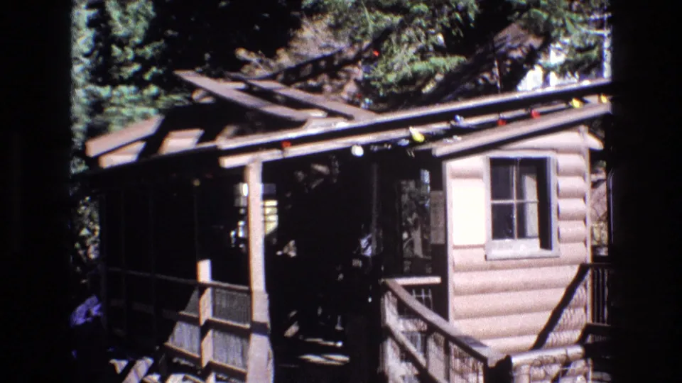 1973 Colorado 8mm frame with the camera closer on a rustic log-style building, porch steps or railings, a window, overhead track supports, colored string lights, and surrounding evergreens.
