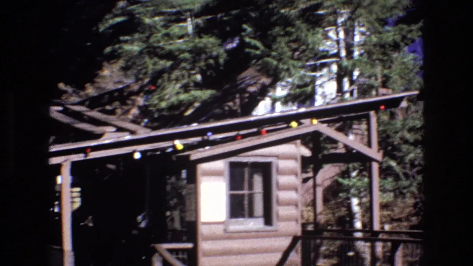 1973 Colorado 8mm frame showing the log-style cabin or station from another angle, with overhead cable supports, porch railing, colored lights, trees, and rocky ground in view.