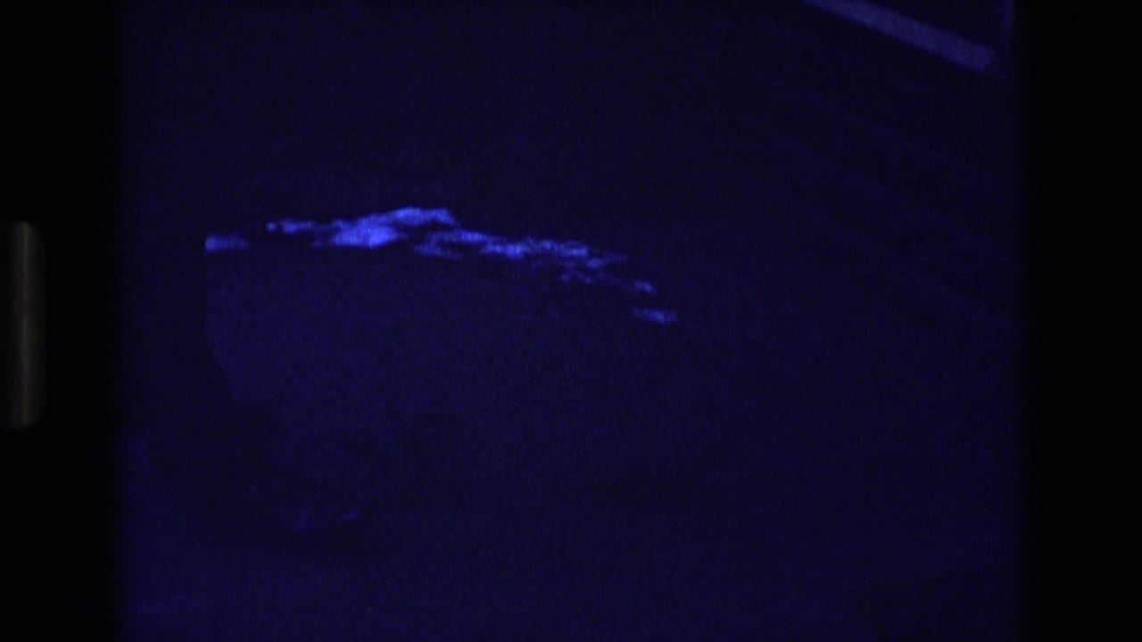 Low-light 1973 Colorado 8mm frame showing a faint hand extended toward a blue-lit patch on the ground, with surrounding details mostly obscured by darkness.
