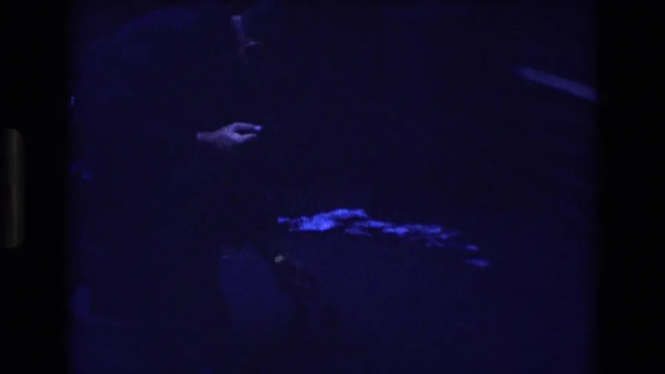 Low-light 1973 Colorado 8mm frame showing a pale hand extended in darkness, with faint bird-like shapes or feeding activity near the lower part of the frame.