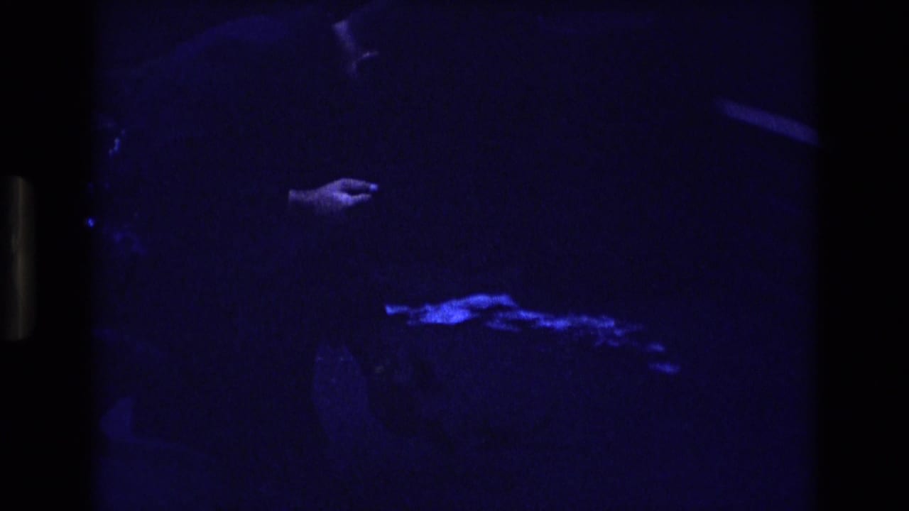 Dark blue-tinted frame with a partial figure and an outstretched hand near a small bright object on the ground.