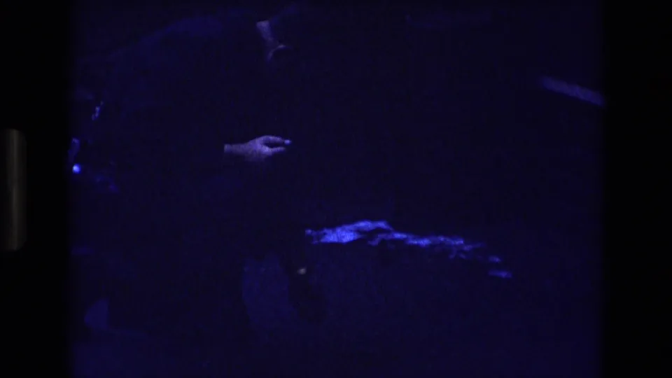 In this dark 8mm frame from the 1973 Colorado clip, a person appears to lean forward while a hand and bright blue patch remain visible near the ground.