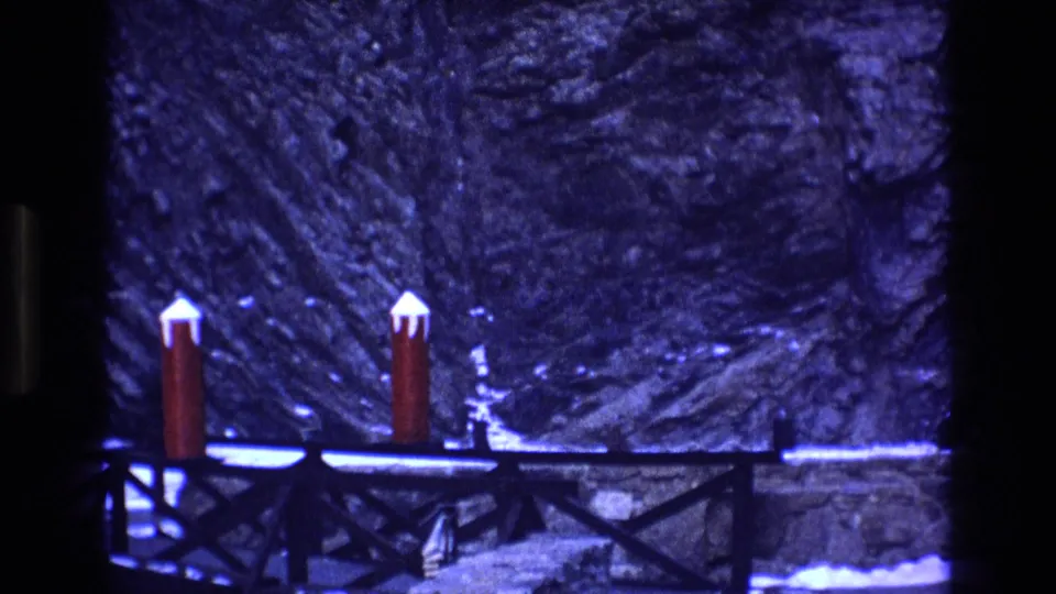 1973 Colorado 8mm frame showing a snowy outdoor bridge area with two red posts, dark wooden rails, and a rocky or wooded background under a strong blue cast.