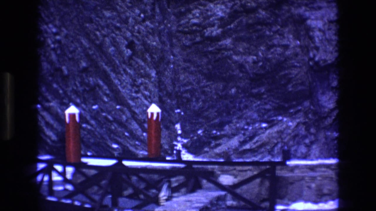 1973 Colorado 8mm frame showing two red posts topped with snow beside a dark bridge or walkway, with a rocky snowy hillside behind them.