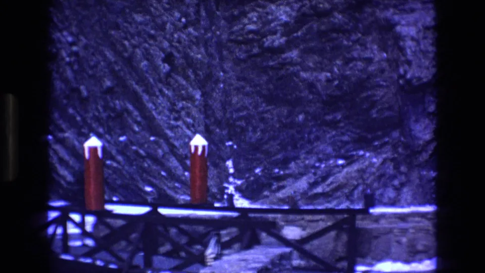 1973 Colorado 8mm frame showing two red posts topped with snow beside a dark bridge or walkway, with a rocky snowy hillside behind them.