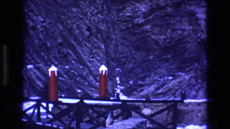 1973 Colorado home-movie frame with red snow-capped posts, a dark bridge railing, and blue-tinted rocky terrain with snow patches.