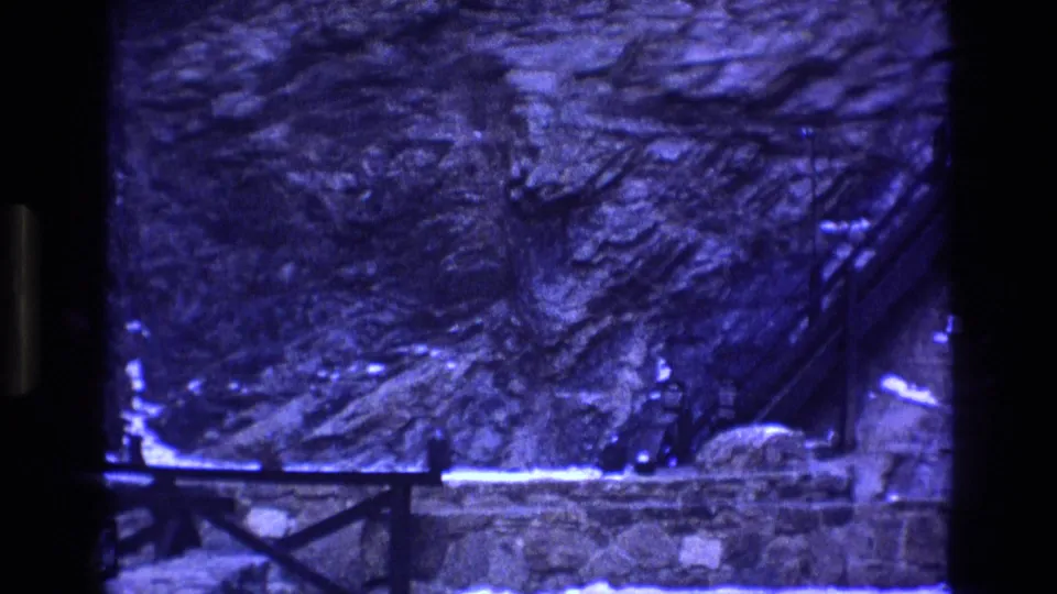 1973 Colorado 8mm frame with a blue-tinted rocky hillside, snow patches, bridge railing at the lower edge, and a dark film-frame border.