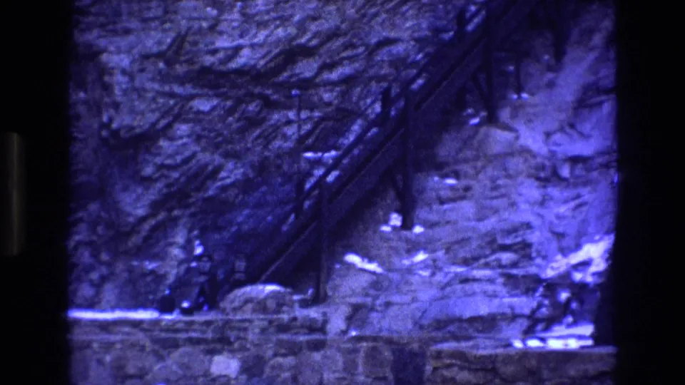 Blue-tinted 1973 Colorado mine footage frame with a steep rock wall, diagonal industrial structure, stonework, and a distant person at the base.