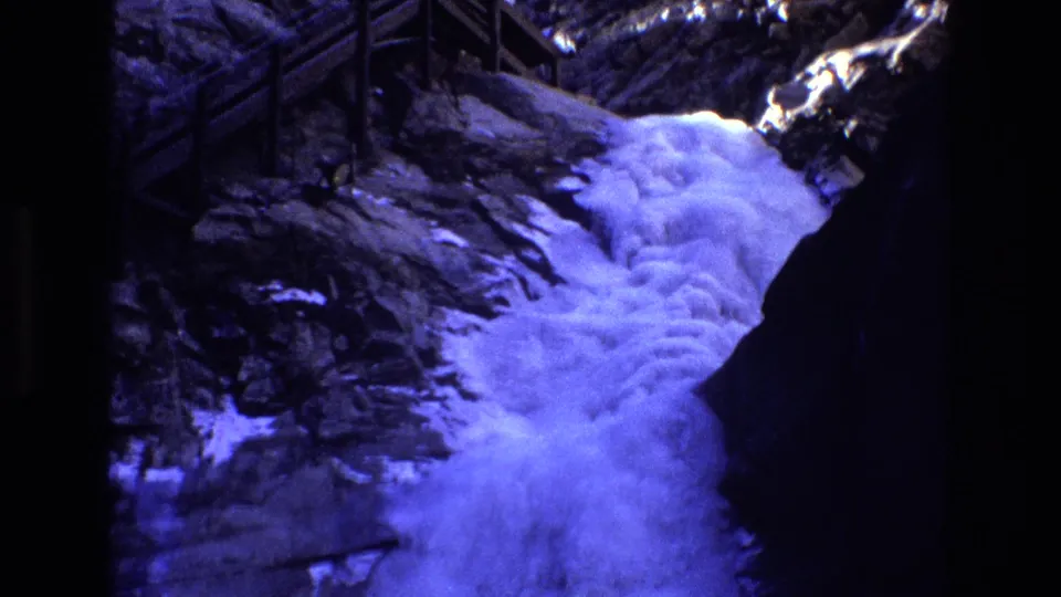 1973 Colorado home-movie frame with blue-purple tinted water pouring through a narrow rocky channel near a raised wooden walkway and railings.