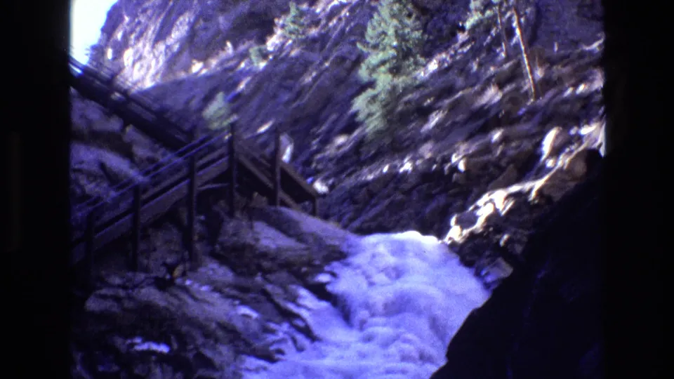 1973 Colorado frame showing turbulent water in a rocky gorge with wooden railings, evergreen trees, bright rock highlights, and saturated blue-purple 8mm color.
