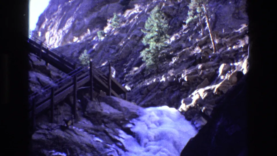 1973 Colorado 8mm frame showing a wooden staircase along a steep rocky canyon wall with foaming water at the base and evergreens on the slope.