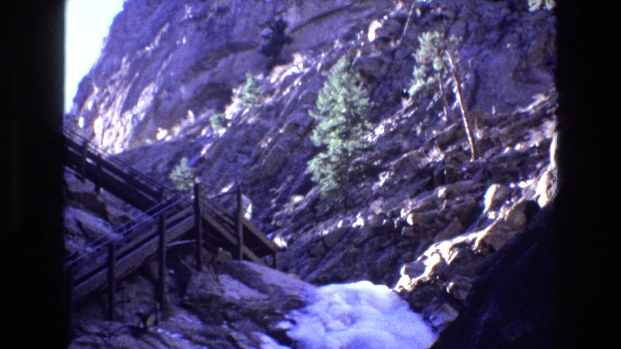 Rocky canyon slope with wooden railings, evergreen trees, and white water below.