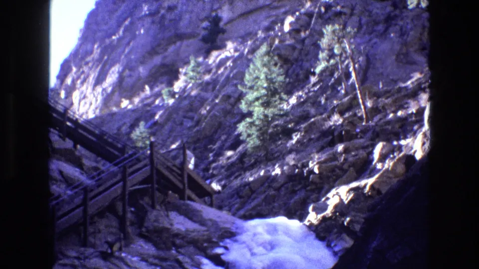 1973 Colorado 8mm frame of a rugged canyon trail where dark wooden railings cross the lower rock face above bright cascading water.