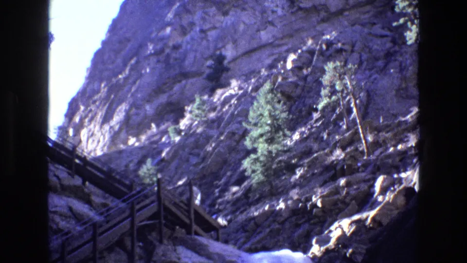 1973 Colorado 8mm frame showing the same mountain canyon from a slightly shifted view, with stairs at left, evergreens on the rocks, and white water below.