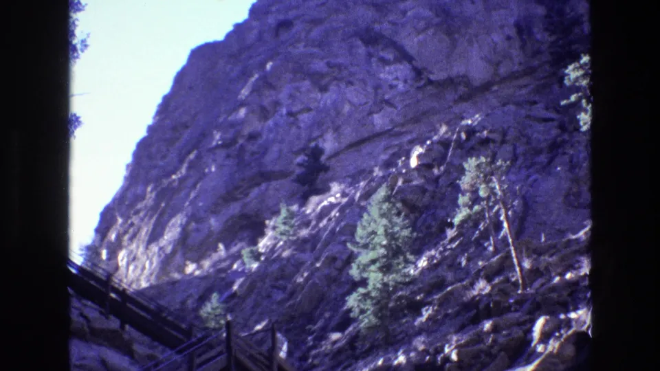 1973 Colorado 8mm frame holding on a steep purple-blue tinted rock wall, a wooden stair rail at lower left, and evergreen growth above the canyon water.