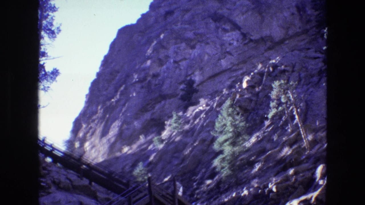 1973 Colorado home-movie frame of a high rocky canyon wall, evergreen growth, and a raised wooden walkway partly silhouetted along the lower edge.