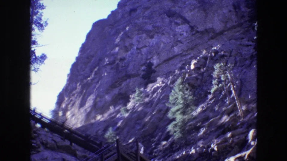 1973 Colorado home-movie frame of a high rocky canyon wall, evergreen growth, and a raised wooden walkway partly silhouetted along the lower edge.
