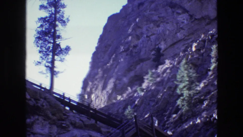 1973 Colorado 8mm frame with a tall evergreen at left, a rocky cliff filling the background, and a wooden bridge or walkway cutting across the lower slope.