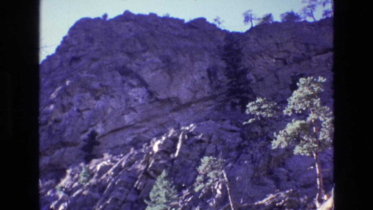 1973 Colorado 8mm frame showing a sunlit rocky cliff face with scattered pine trees and dark film-gate borders.