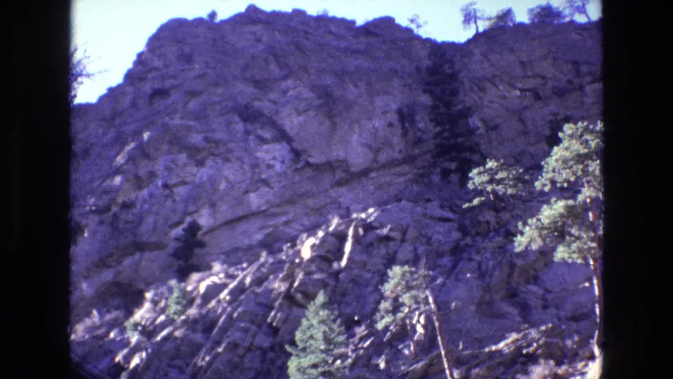 1973 Colorado home-movie frame of a rugged rock wall with small pine trees and high-contrast daylight exposure.