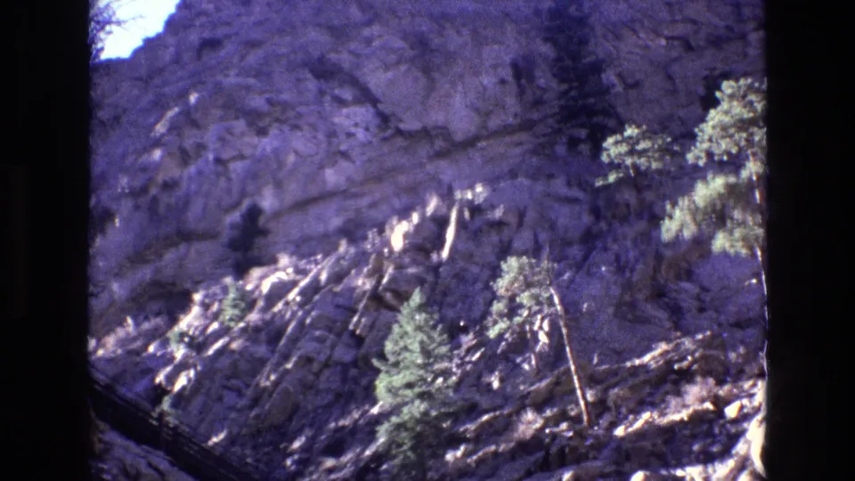 1973 Colorado 8mm frame showing a rocky mountainside, evergreen trees, and a partial dark foreground edge from the film gate.