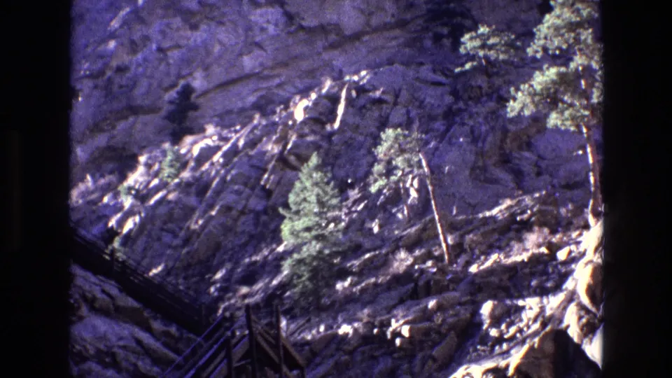 1973 Colorado home-movie frame of a rugged cliff face with pine trees and what appears to be a wooden railing or walkway along the mountainside.