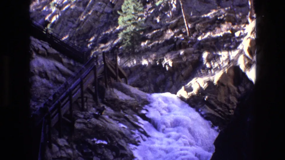 1973 Colorado 8mm frame showing a visitor stairway descending along rugged rock, with foaming waterfall water filling the lower canyon.