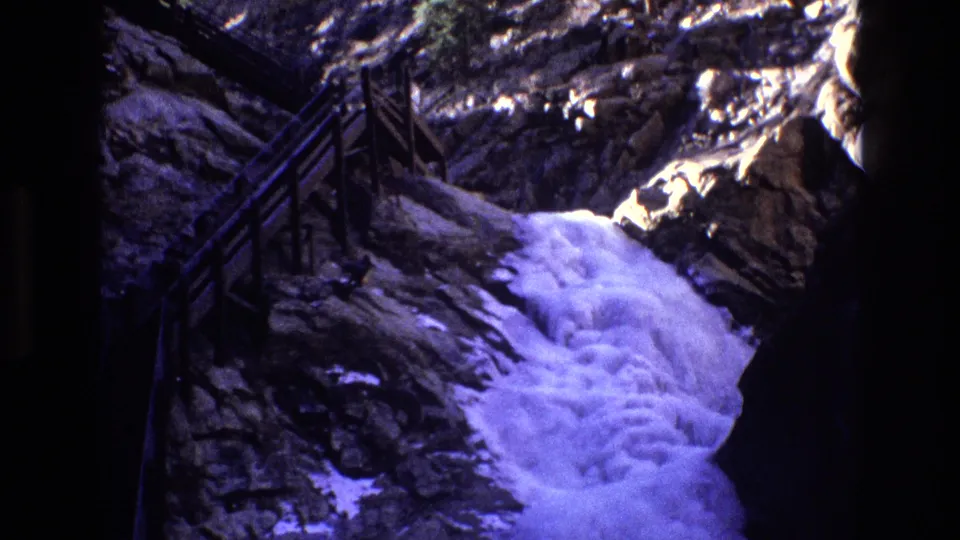 1973 Colorado home-movie frame with bright rushing water at the base of dark rock walls and a wooden stair railing along the canyon side.
