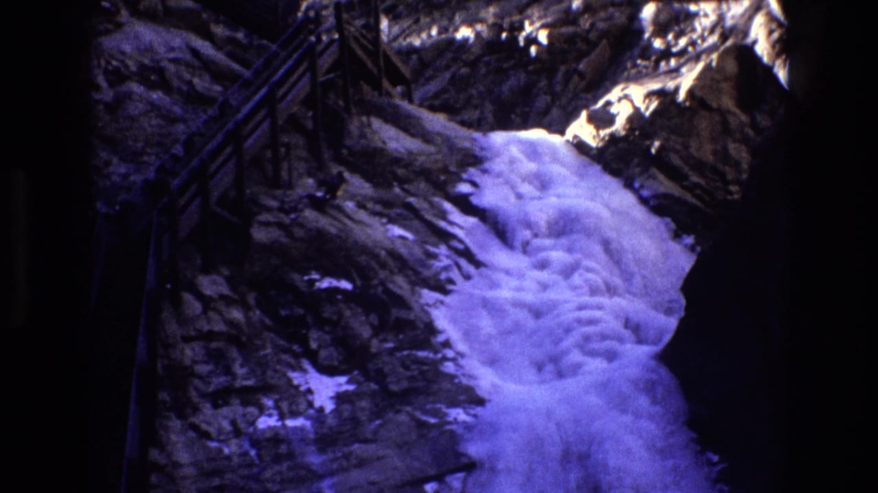 Foaming waterfall runs through dark rock with snow patches and a railing at left.