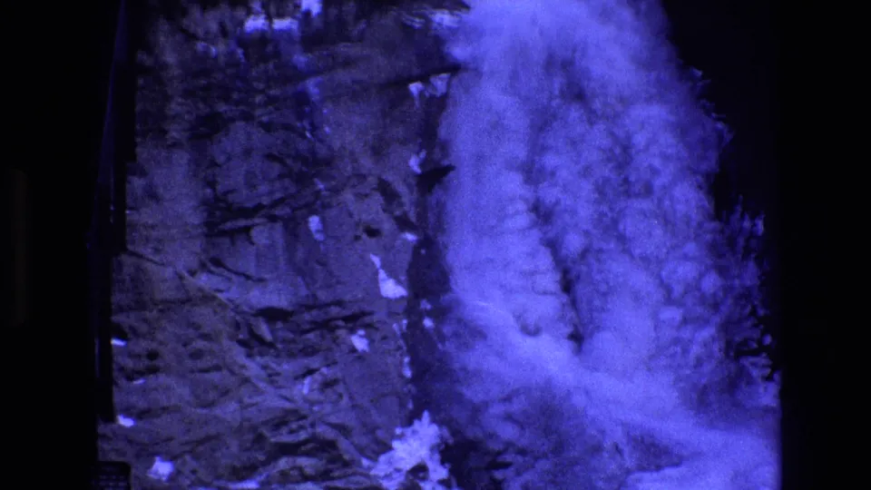 1973 Colorado Super 8 frame showing blue-white water plunging through a narrow dark rock channel with small snow patches visible near the falls.